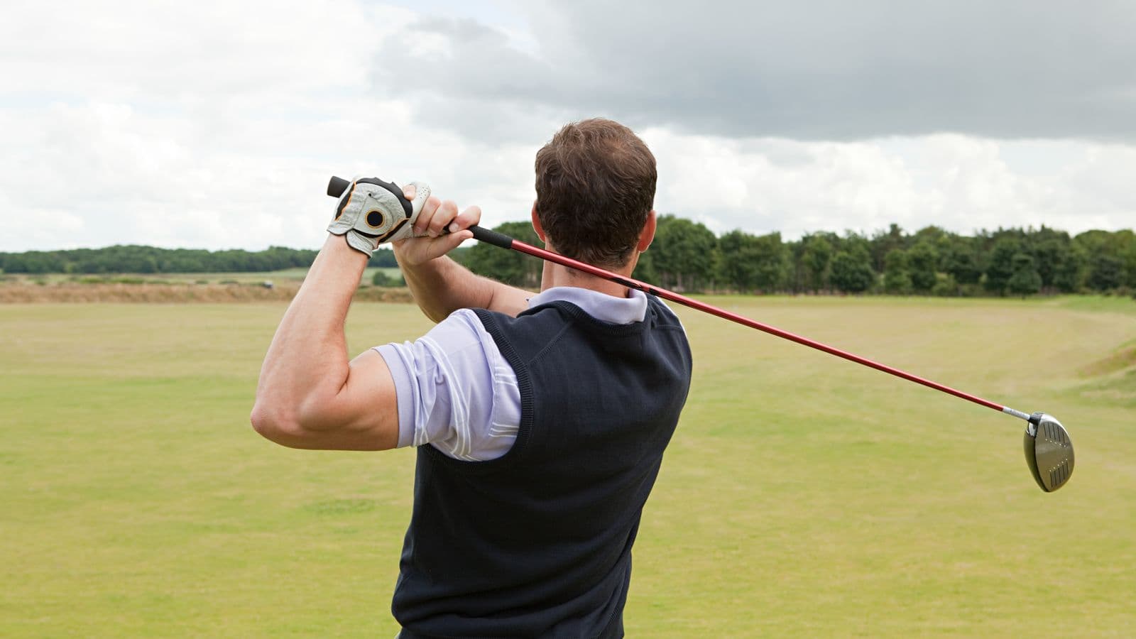 A golfer seen from behind at the top of his backswing with a driver, on an open course under a cloudy sky