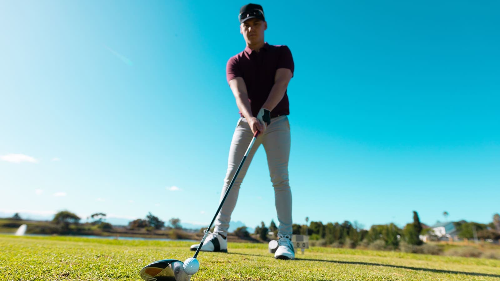 A young golfer in a maroon polo and black cap lines up a teed ball with his driver, seen from ground level under a clear blue sky.