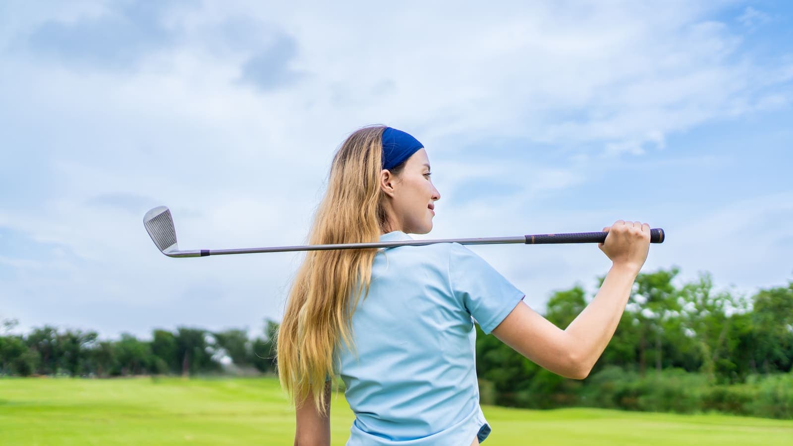 A young woman with long blonde hair and a blue headband rests an iron across her shoulders and looks out over a golf course.