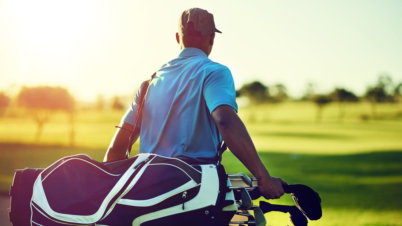 A golfer seen from behind carrying his bag across a fairway in low evening sun