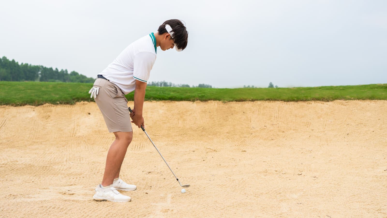 A young golfer in a white polo and visor sets up to play out of a greenside bunker on an overcast day.