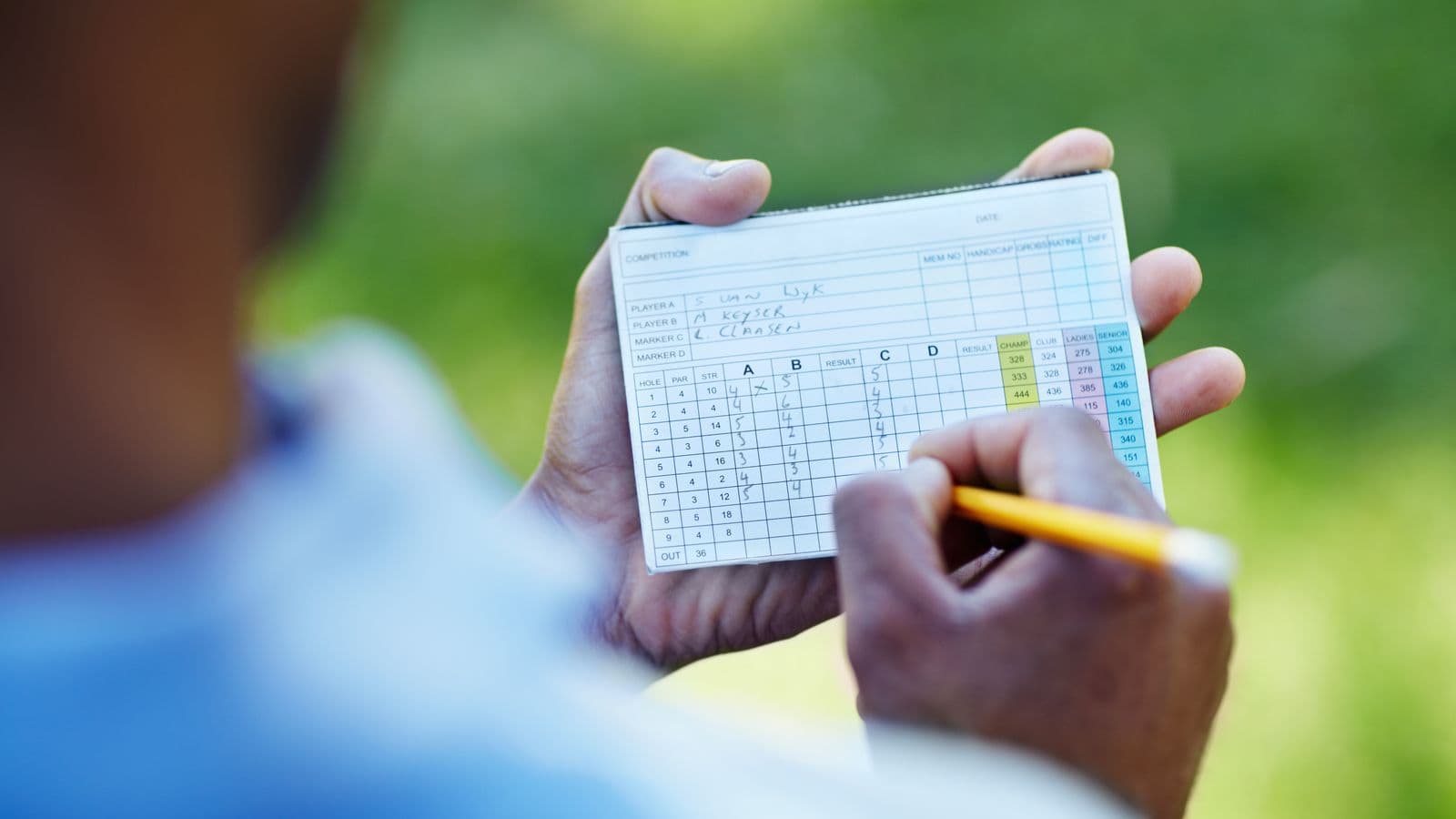 A golfer's hands filling in a scorecard with a yellow pencil on the course