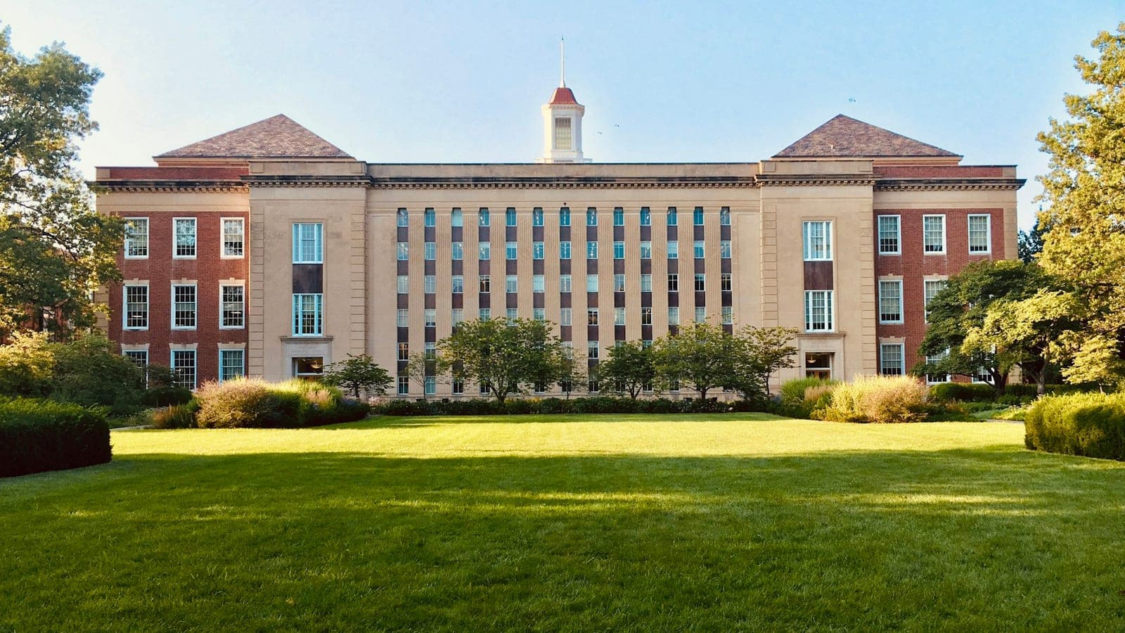 A brick and limestone college building behind a wide green lawn on a clear morning