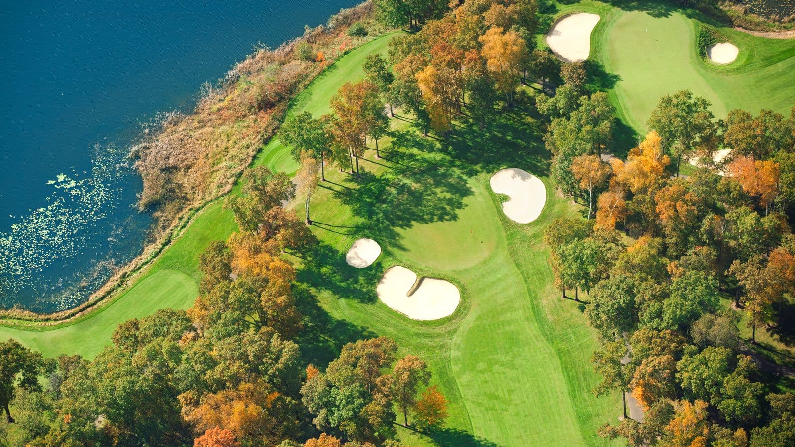 An aerial view of a golf hole in autumn, with sand bunkers along a fairway lined with orange and green trees beside a lake